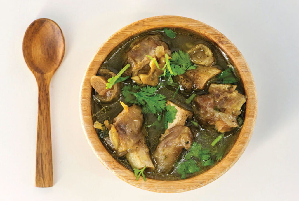 A wooden spoon and bowl of Goat Paya decorated with coriander leaves.