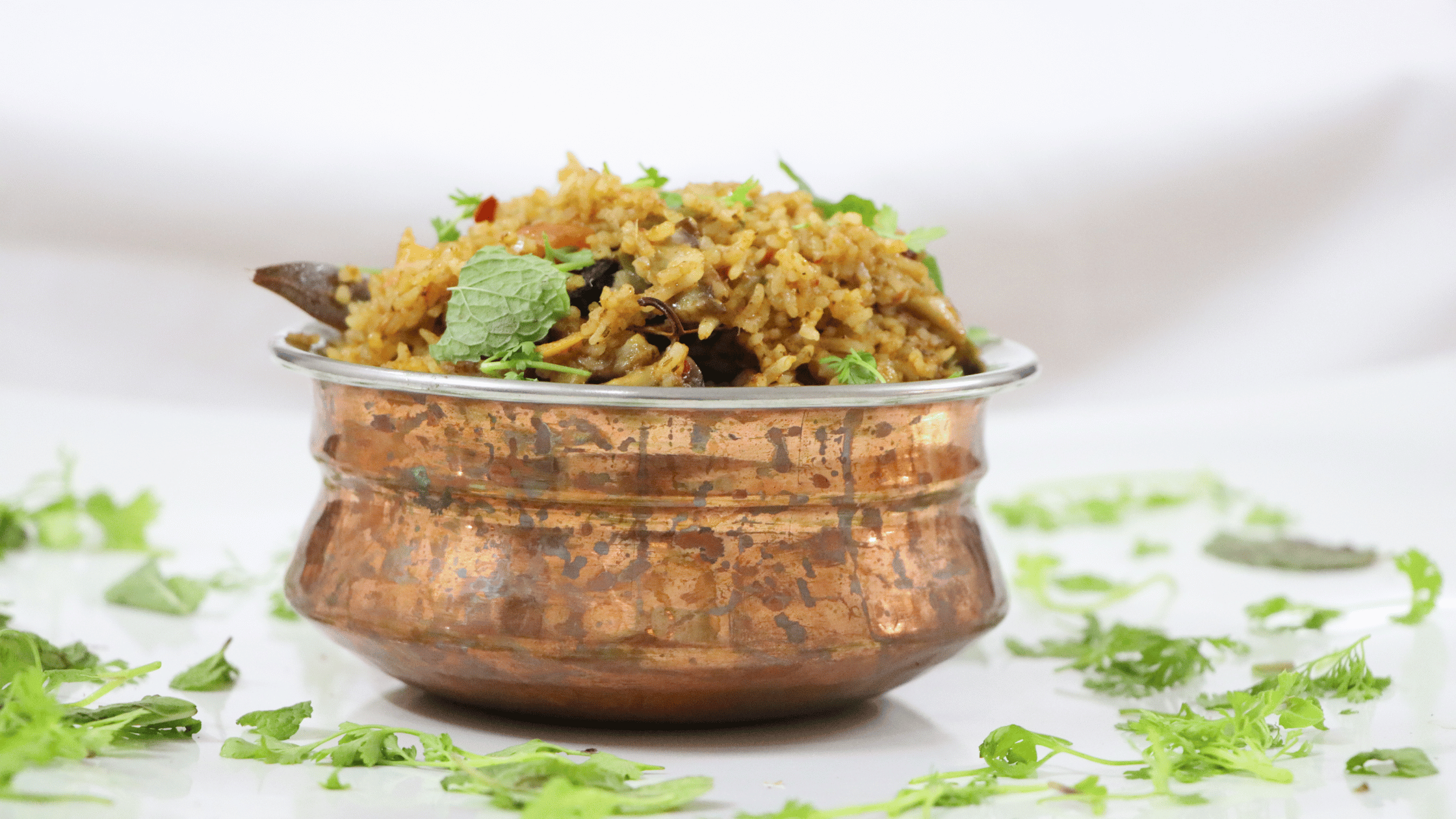 Aromatic biryani served in a traditional bronze container, garnished with fresh herbs and accompanied by sides, showcasing a rich and flavorful presentation.