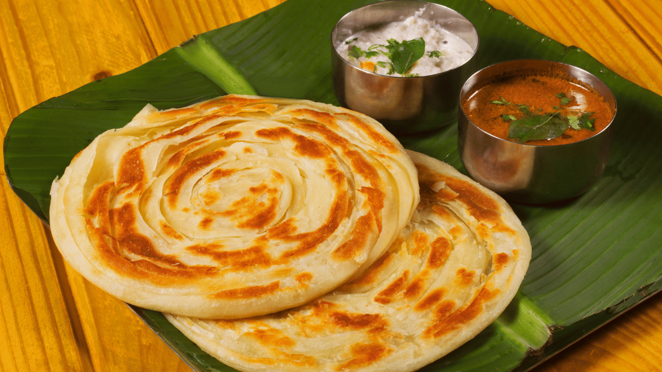 Delicious South Indian parotta served on a banana leaf with flavorful curry and curd raita, presented on a traditional wooden table.