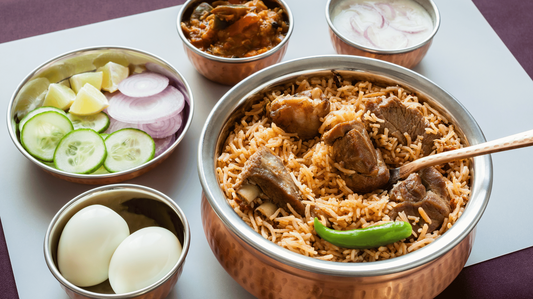 Delicious mutton biryani served in a traditional copper pot with side dishes including boiled eggs, sliced cucumbers, onions, and yogurt raita.