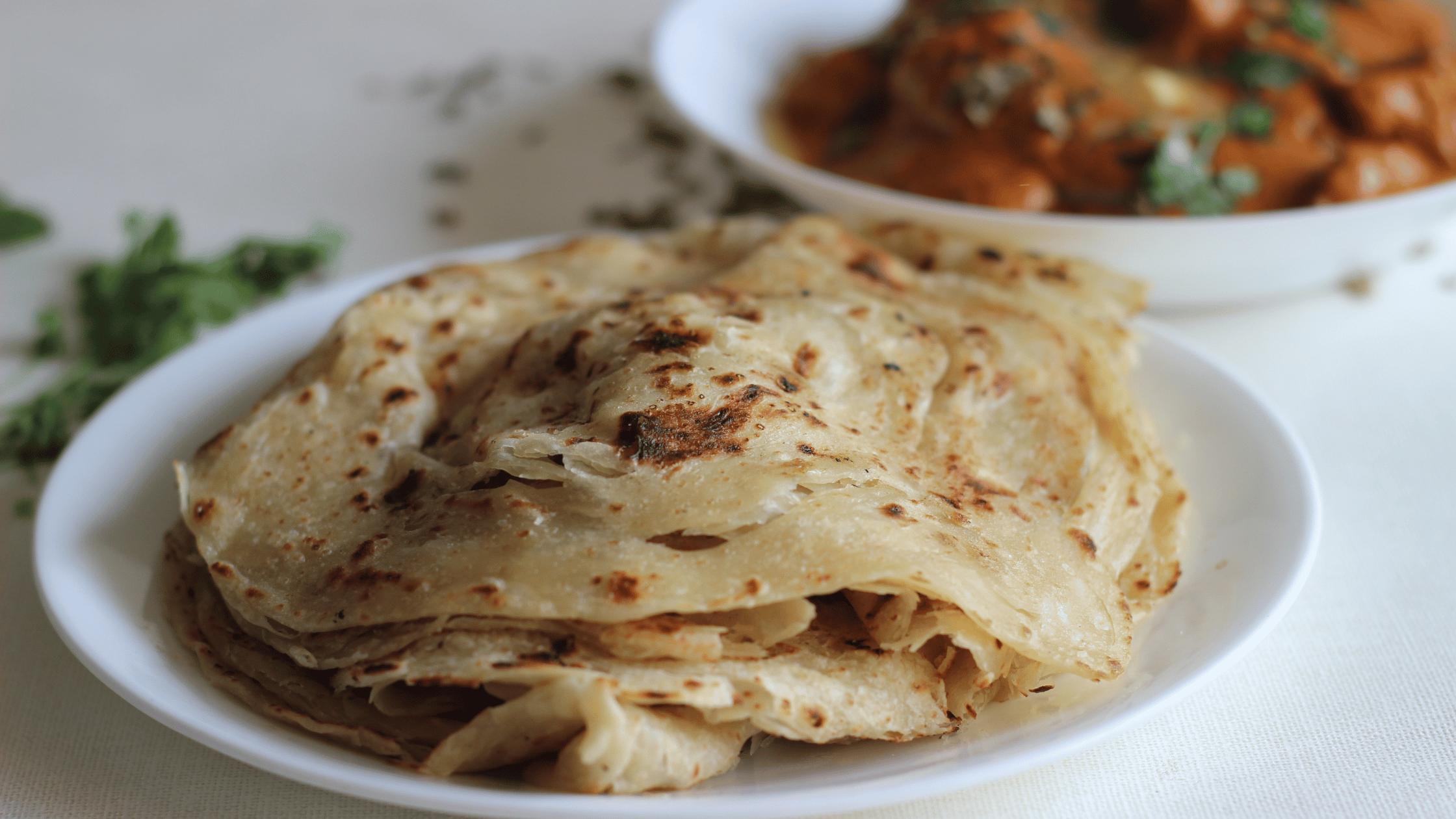 Delicious, flaky South Indian parotta served on a white plate, accompanied by a flavorful curry in the background, highlighting an authentic Indian dining experience.