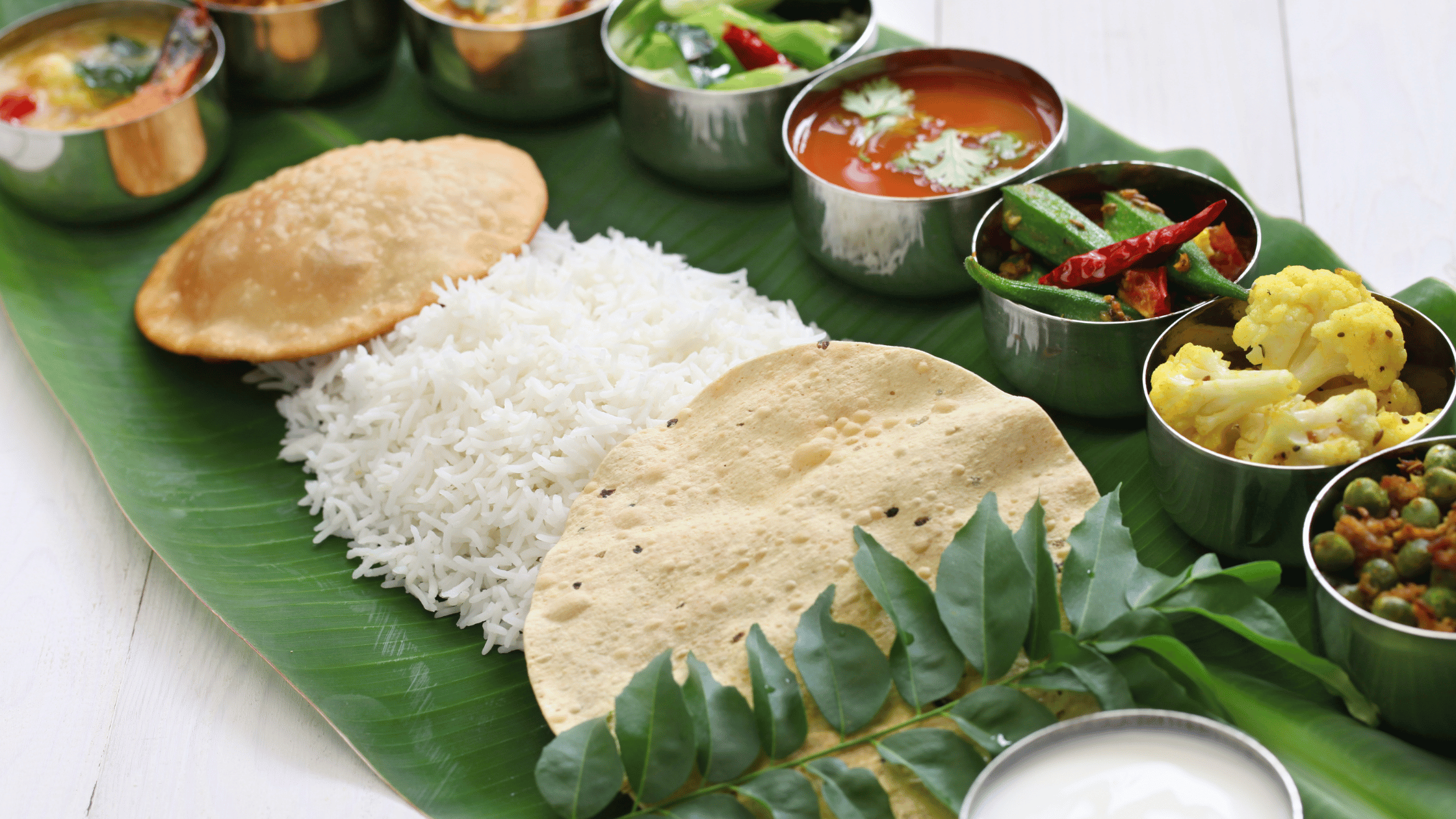 Traditional South Indian meal served on a banana leaf with rice, puri, papad, and a variety of vibrant vegetable curries and side dishes in stainless steel bowls.